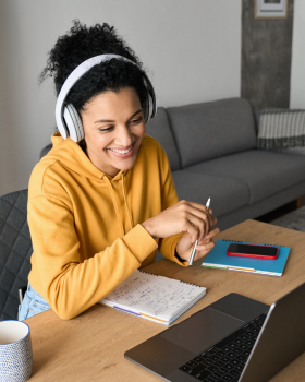 a person sitting at a desk holding a pen wearing headphones and smiling at an open laptop